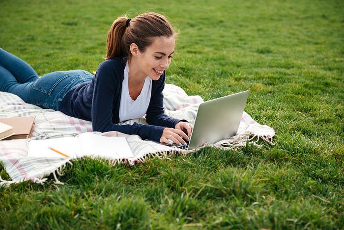 Happy teenage girl laying on blanket outside on the grass, typing on a laptop.